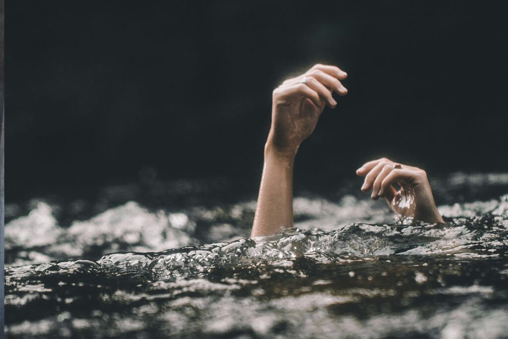 A woman’s hands emerge from a body of water, signifying she is drowning.