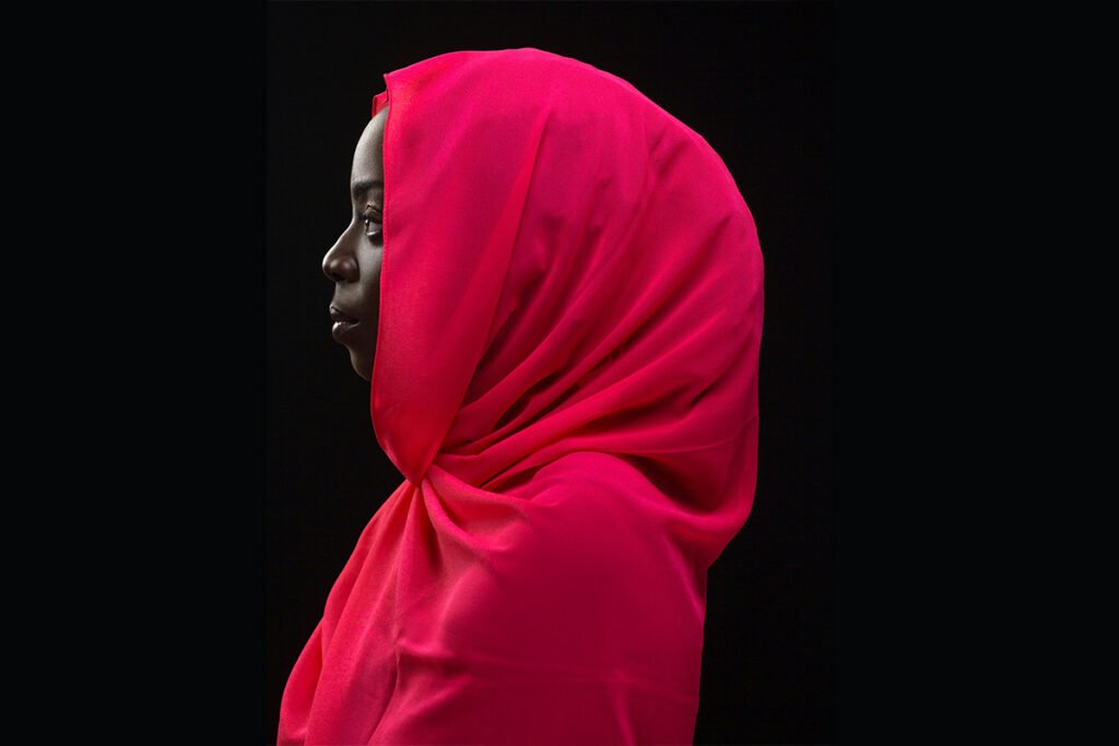 A deep skinned Black woman wears a red hijab and turns in profile to the left against a dark background.