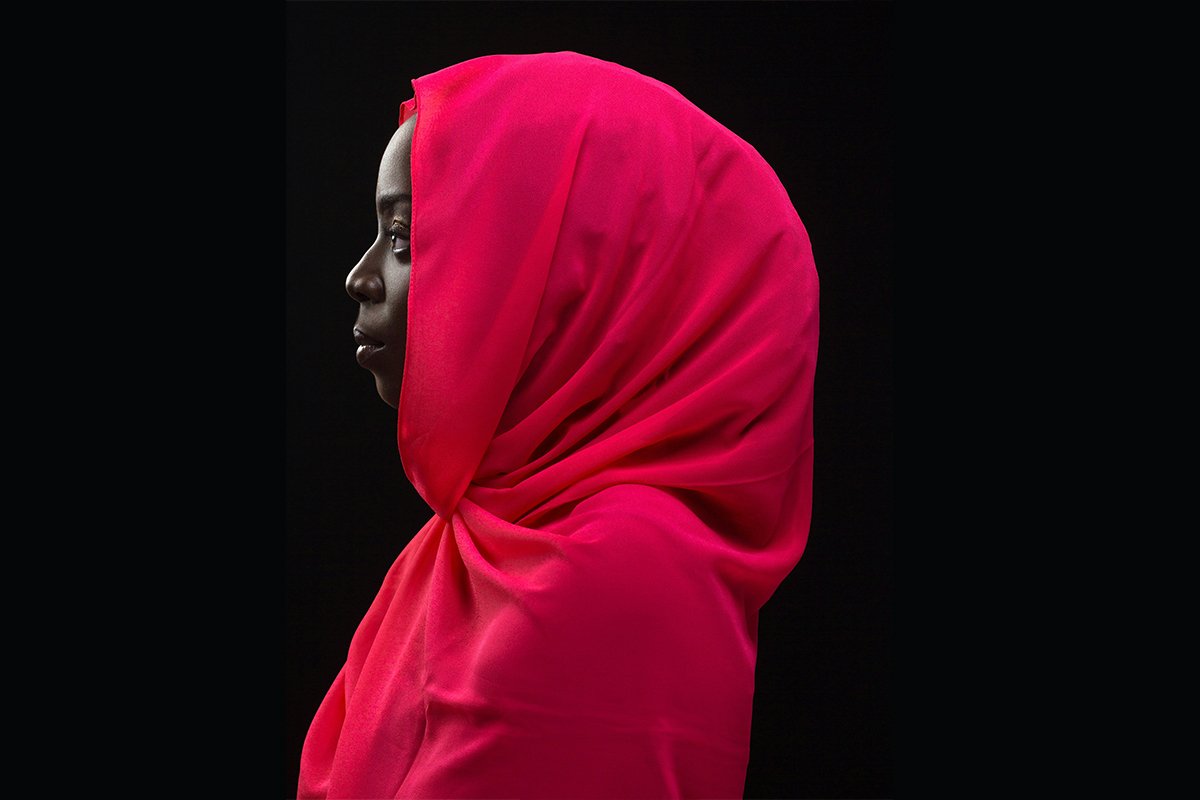 A deep skinned Black woman wears a red hijab and turns in profile to the left against a dark background.