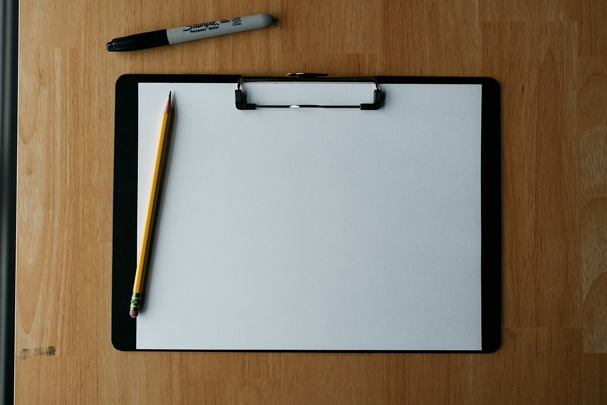 A blank clipboard placed on a wooden table with a pencil and sharpie nearby.