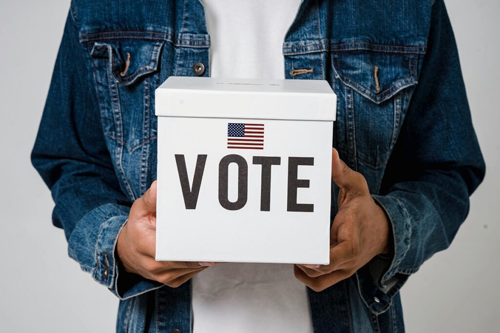 A person in a jean jacket holds up a ballot box with an American flag and the word “vote” on it.