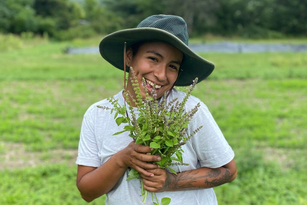 A smiling brown-skinned person wearing a sun hat and hold a tuft of plants. They are standing in a field of grass.