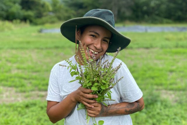 A smiling brown-skinned person wearing a sun hat and hold a tuft of plants. They are standing in a field of grass.