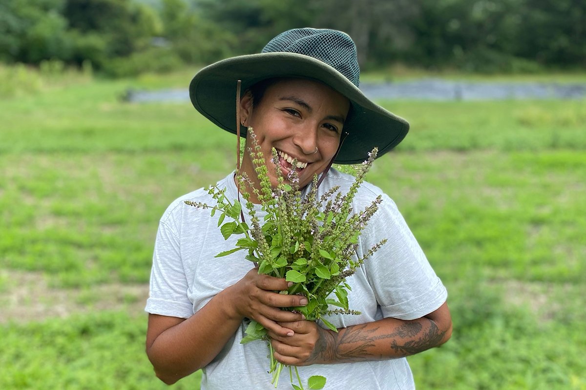 A smiling brown-skinned person wearing a sun hat and hold a tuft of plants. They are standing in a field of grass.