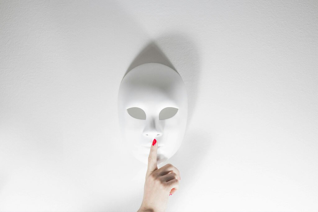 A white mask lays against a white background. A human hand holds up a pointer finger in front of the mask, in the “quiet” pose.