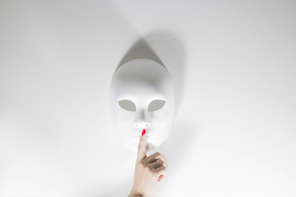 A white mask lays against a white background. A human hand holds up a pointer finger in front of the mask, in the “quiet” pose.