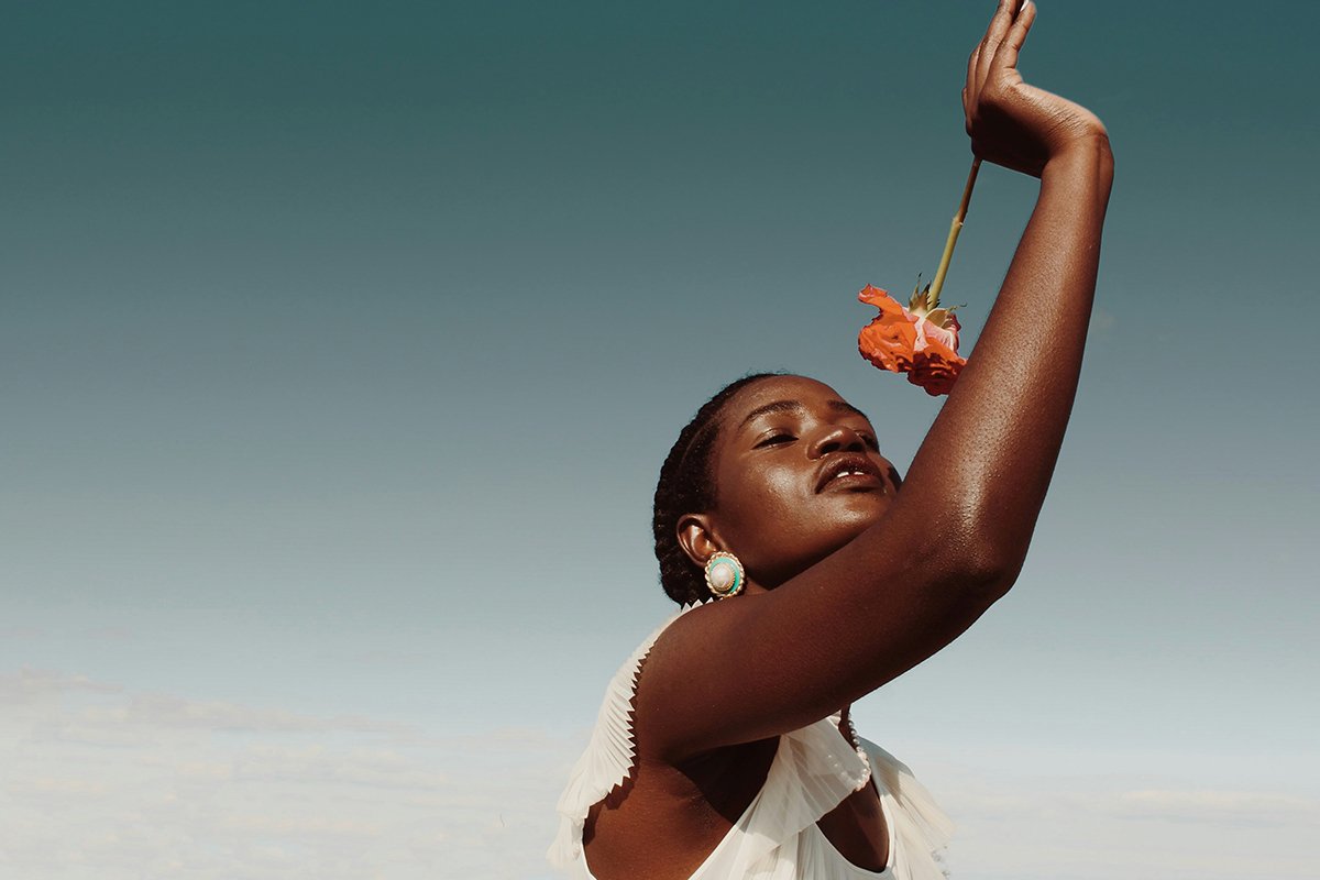 A Black woman with cornrows and a flowy white shirt holds a red rose up to her face as she poses against a cloudless sky.
