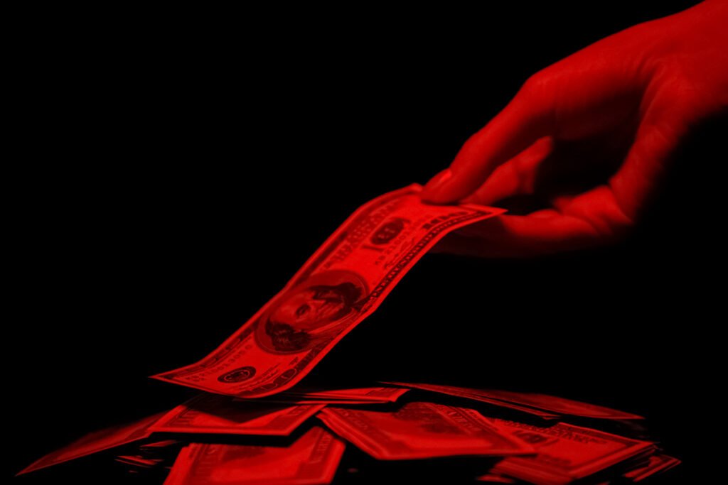 A hand takes a dollar bill of the top of a pile of bills. The scene is washed in red, ominous light.