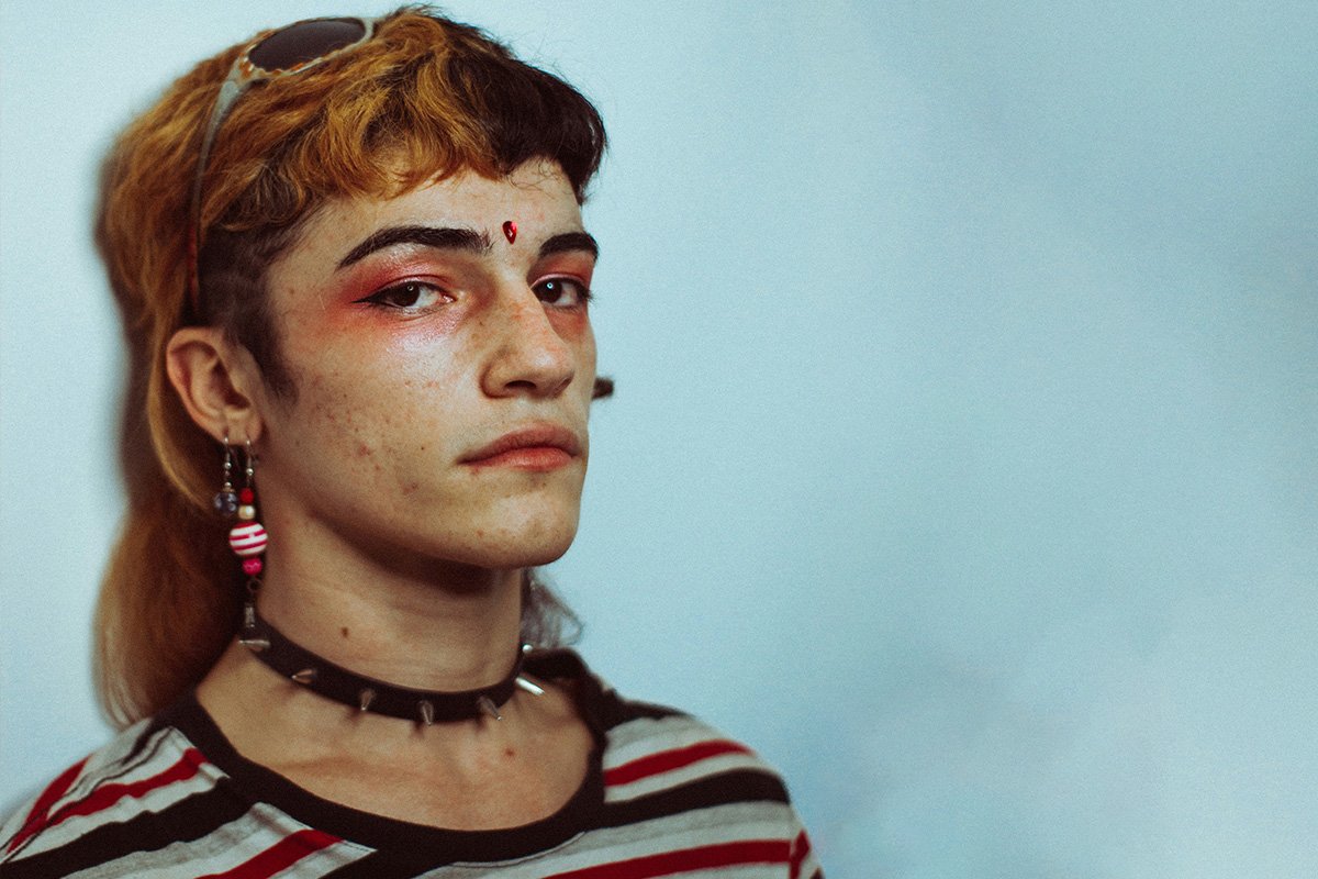 An androgynous individual with a split-dyed mullet and pink eyeshadow looks into the camera sullenly.