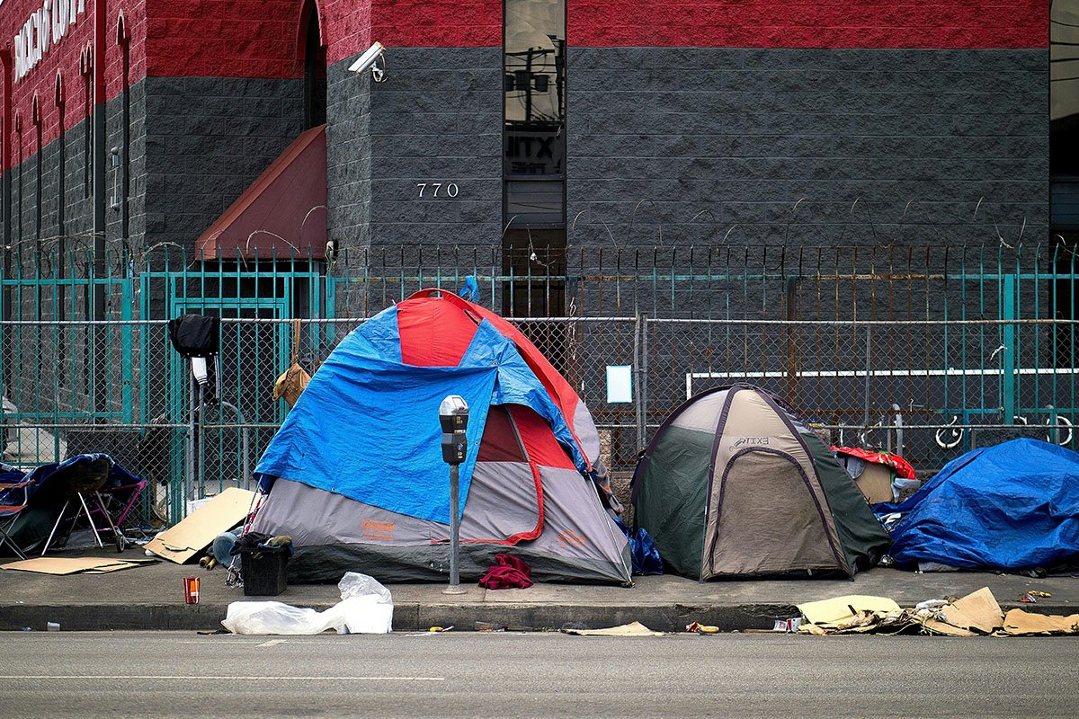 An unhoused encampment in Los Angeles, CA with a blue and red tent next two two smaller tents, on a sidewalk with a chain link fence.