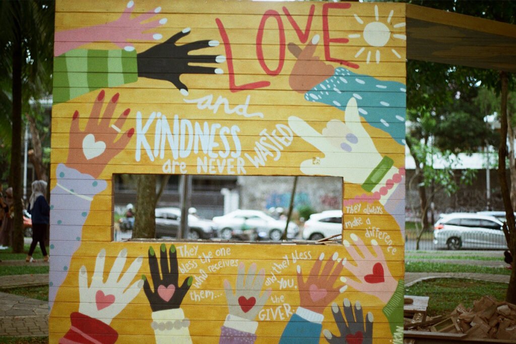 A painted wooden wall with many-colored hands and hearts, with the words, “Love and Kindness are never wasted”