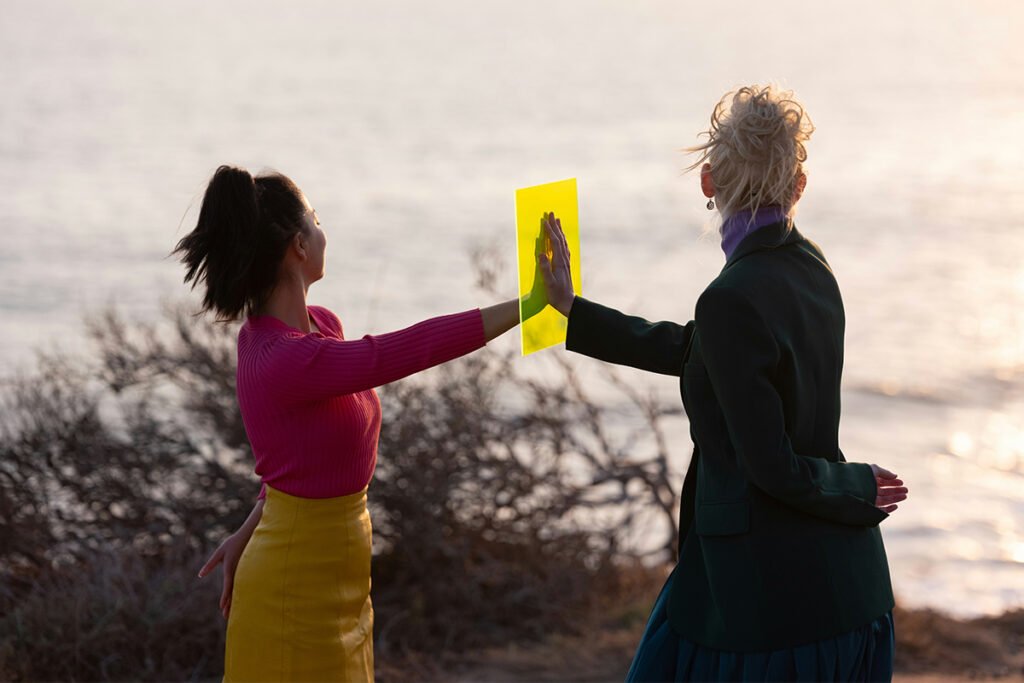Two women stand in front of each other and face away from the camera. Between them, they press their hands together against a yellow frame.