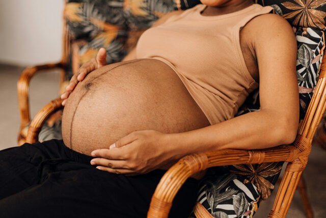 A Black woman sits in a tropical-patterned lounger cradling her belly in her hands.