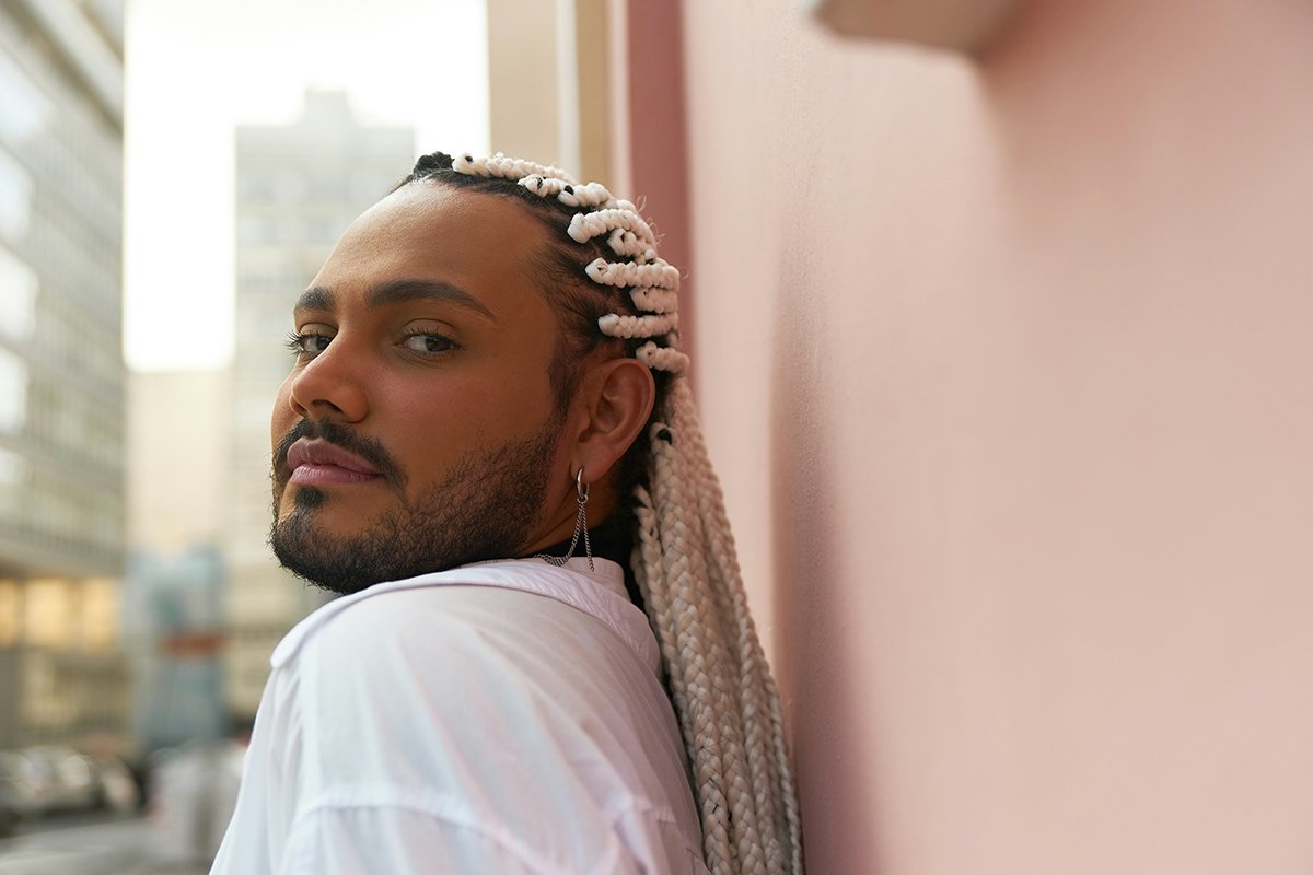 A person with a goatee and long platinum braids looks over their shoulder and into the camera knowingly. The background is a cityscape.
