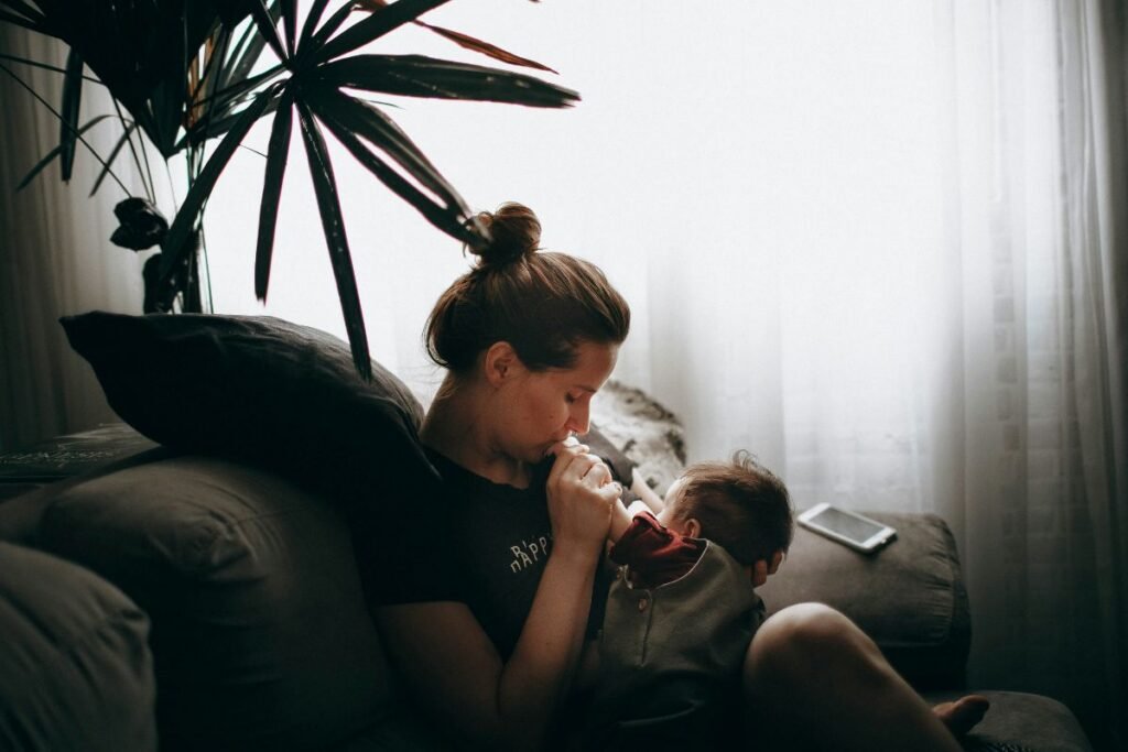 A woman sits on a couch, holding an infant with dark hair and kissing its hand. A houseplant and closed curtain are in the background.
