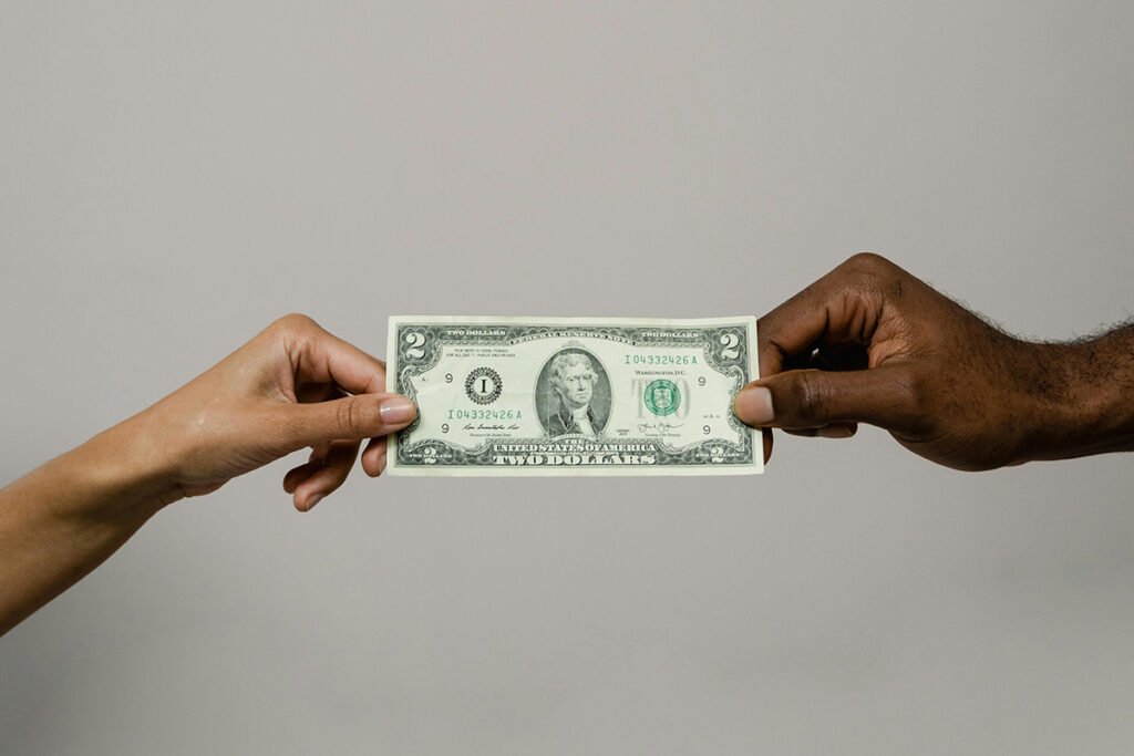 A white hand and a Black hand hold either side of a rare, two dollar bill.