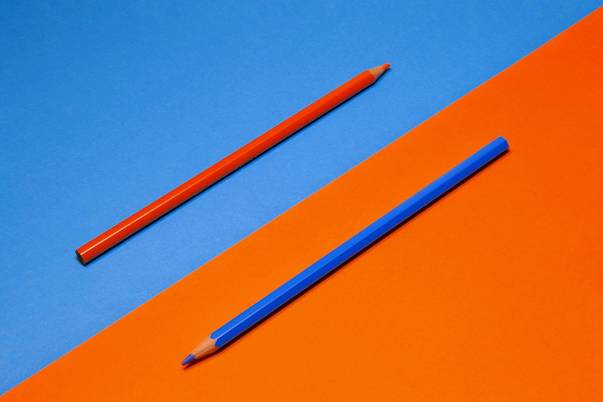 Two colored pencils–orange and blue–placed on a split background. The orange pencil is on a blue background and the blue pencil is on an orange background.