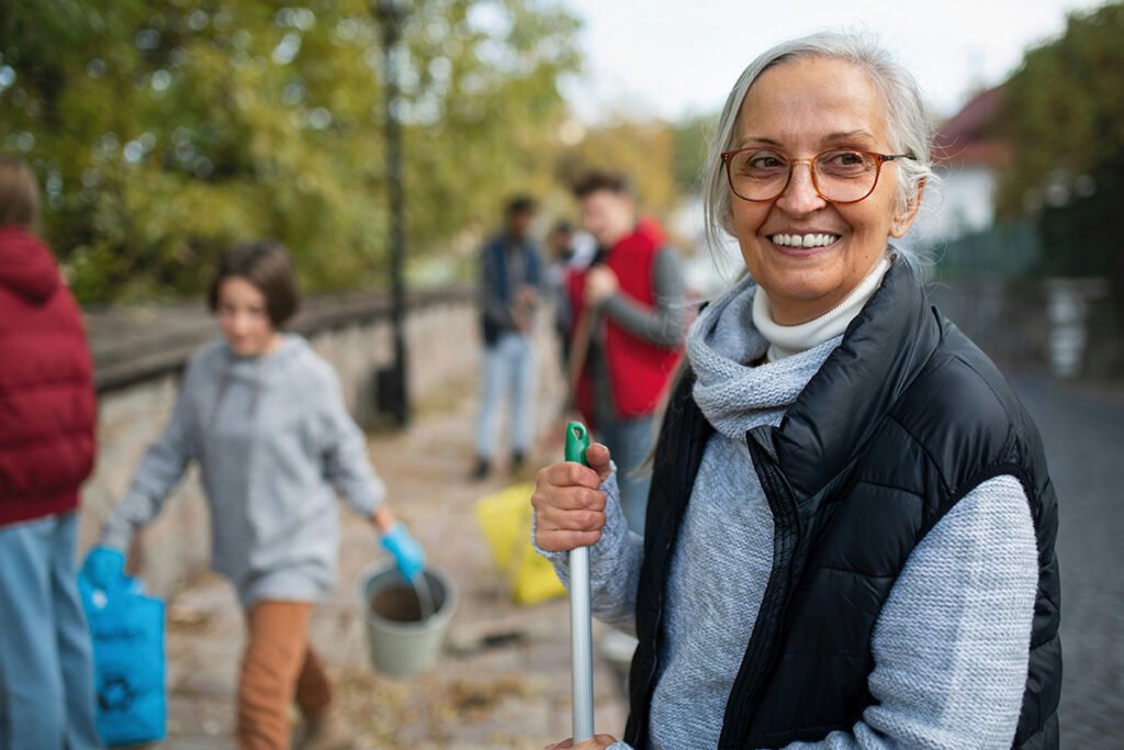 An elder woman with long gray hair and a black vest volunteers outside, holding a tool in one hand while other volunteers walk around behind her.
