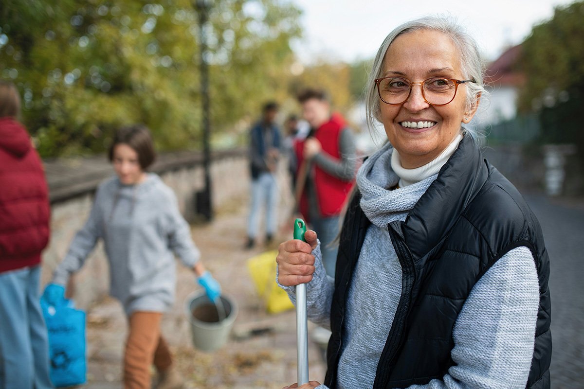 An elder woman with long gray hair and a black vest volunteers outside, holding a tool in one hand while other volunteers walk around behind her.