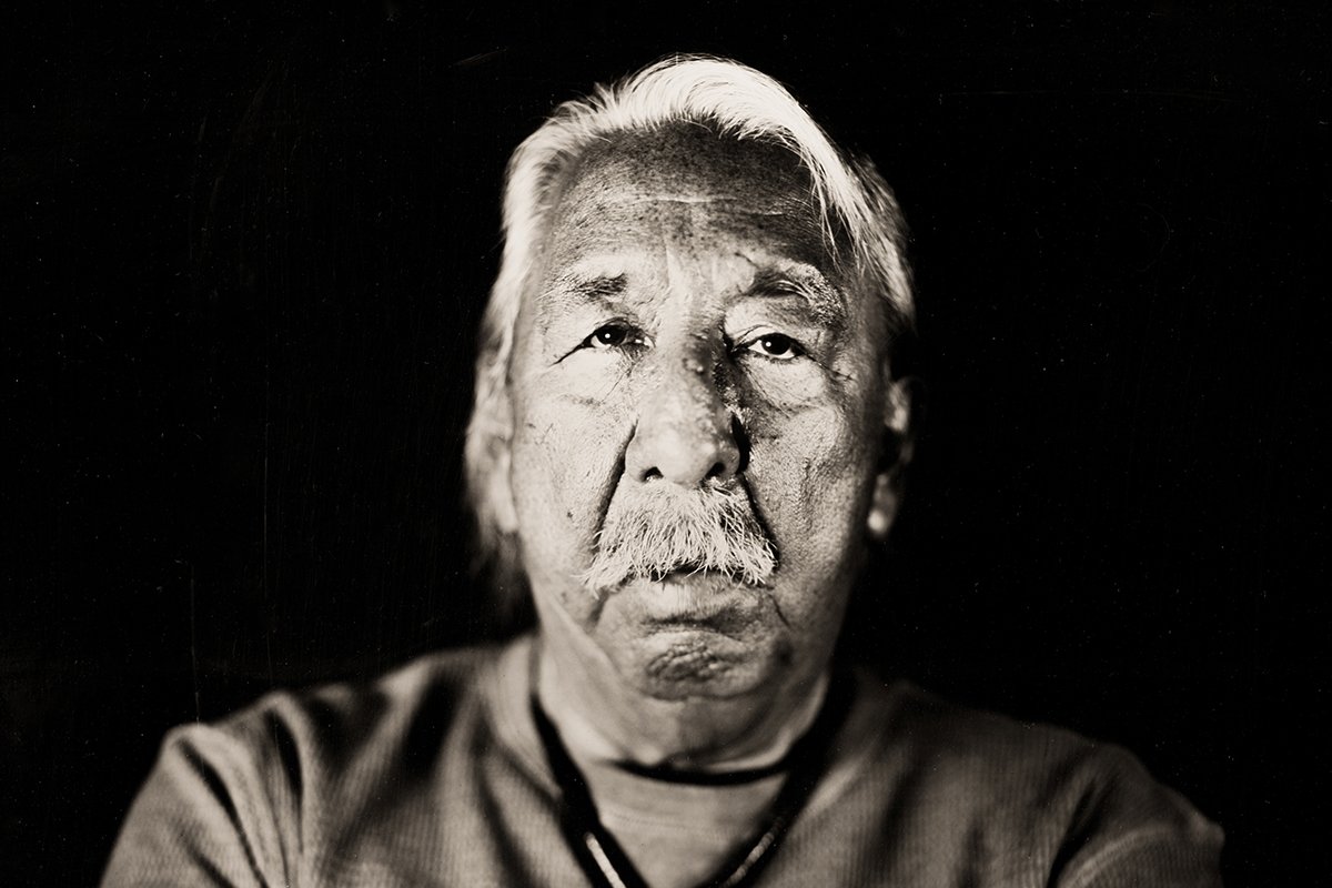 Leonard Peltier (Chippewa / Lakota) photographed in Black and white looking directly at the camera. He is an older man with sun-spotted skin and medium length white hair swept to the side.