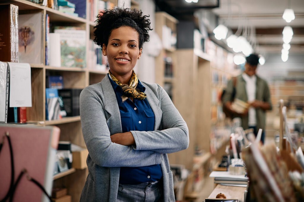 A Black woman with natural hair stands behind the check-out counter at a bookstore. The shelves behind her are lined with books.
