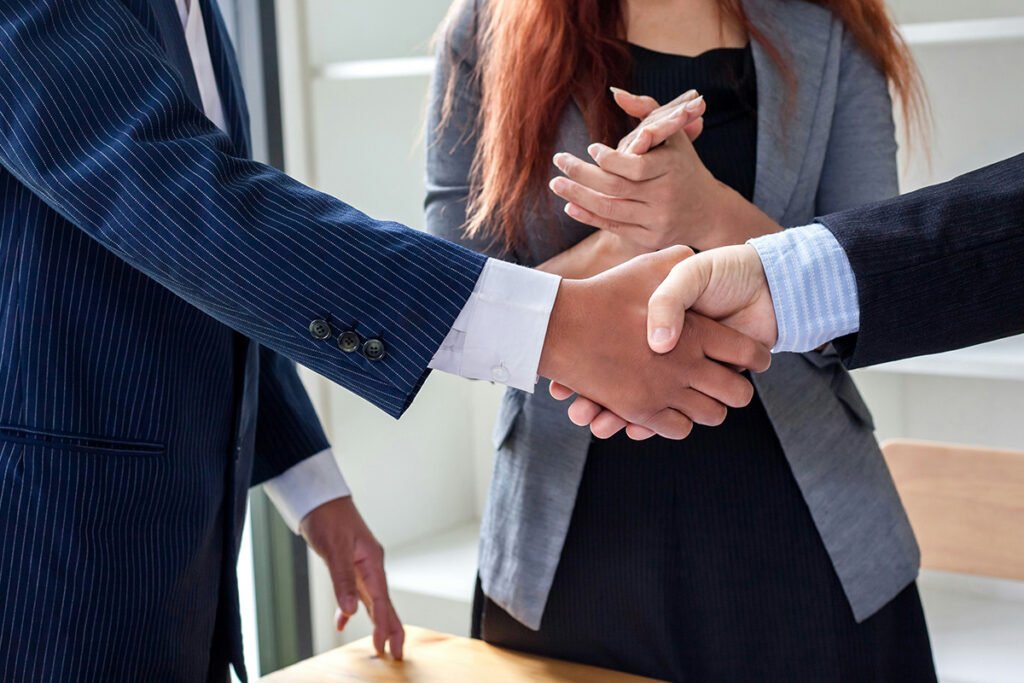 Two people in suits shake hands as a professionally-dressed woman looks on and clasps her hands during a professional introduction.
