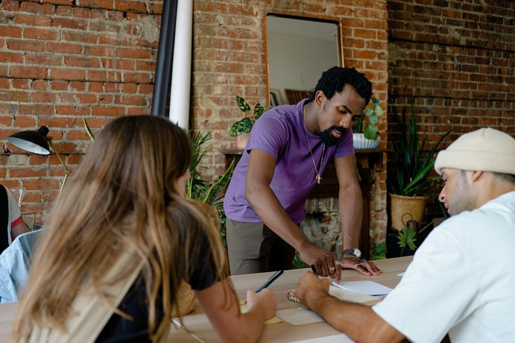A small, but diverse group of people join each other at a table and workshop together while holding papers and notepads.
