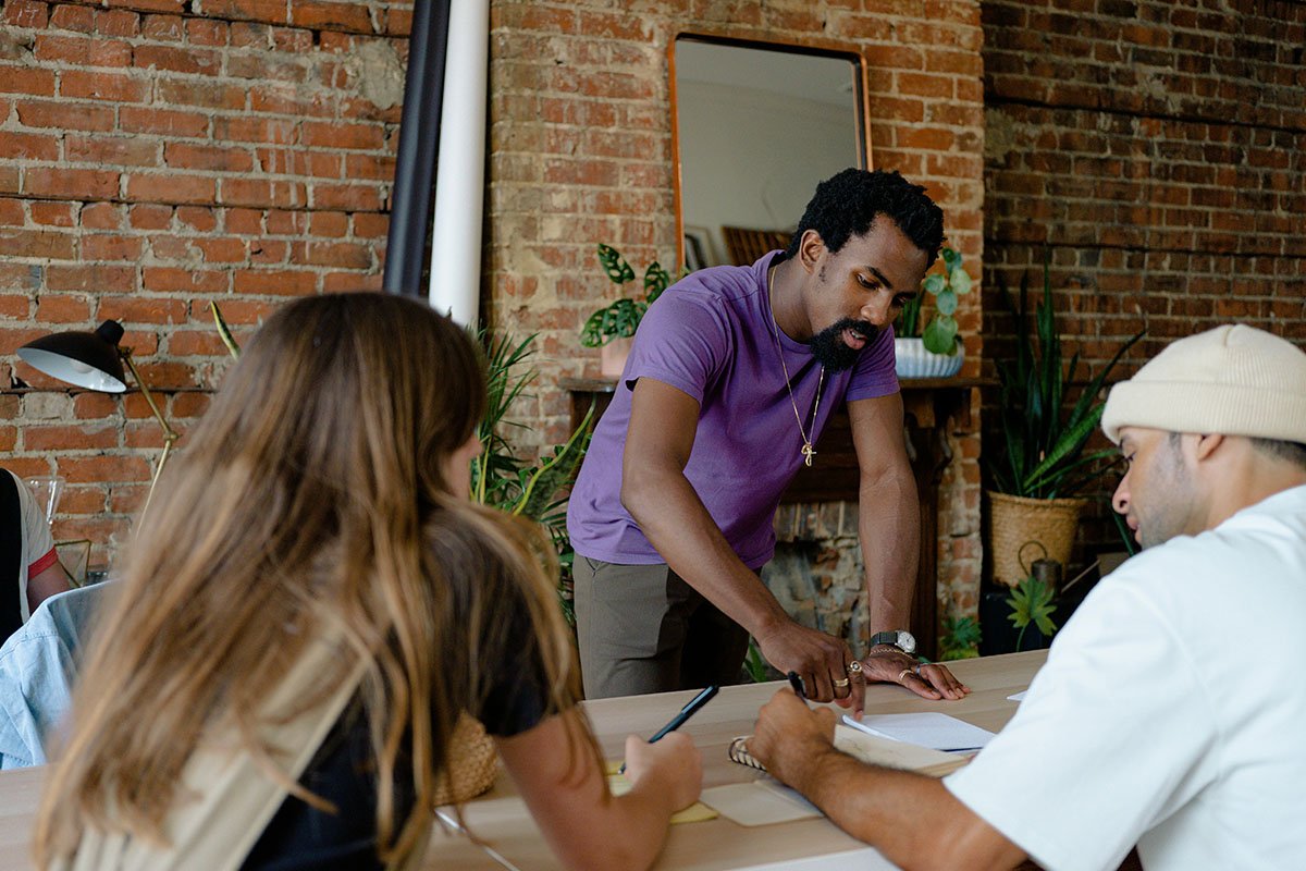 A small, but diverse group of people join each other at a table and workshop together while holding papers and notepads.