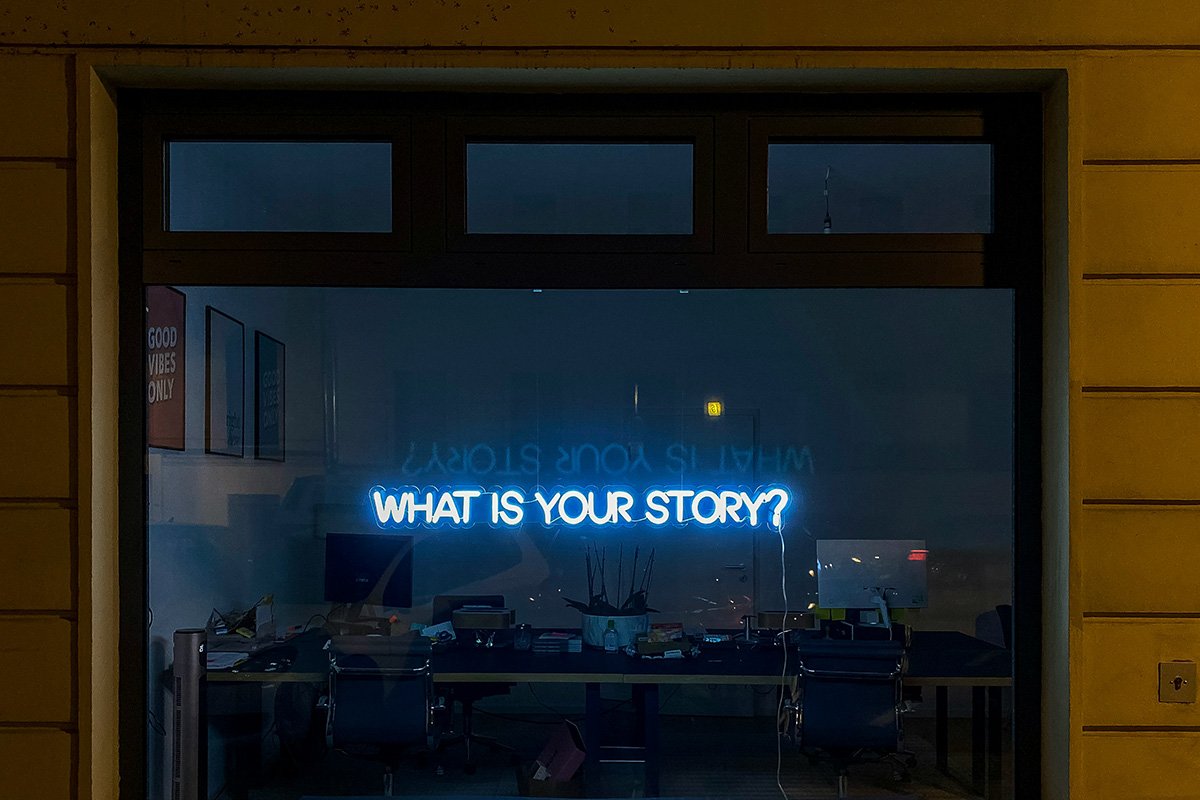 A blue neon sign in a store window that reads, “What is Your Story?”