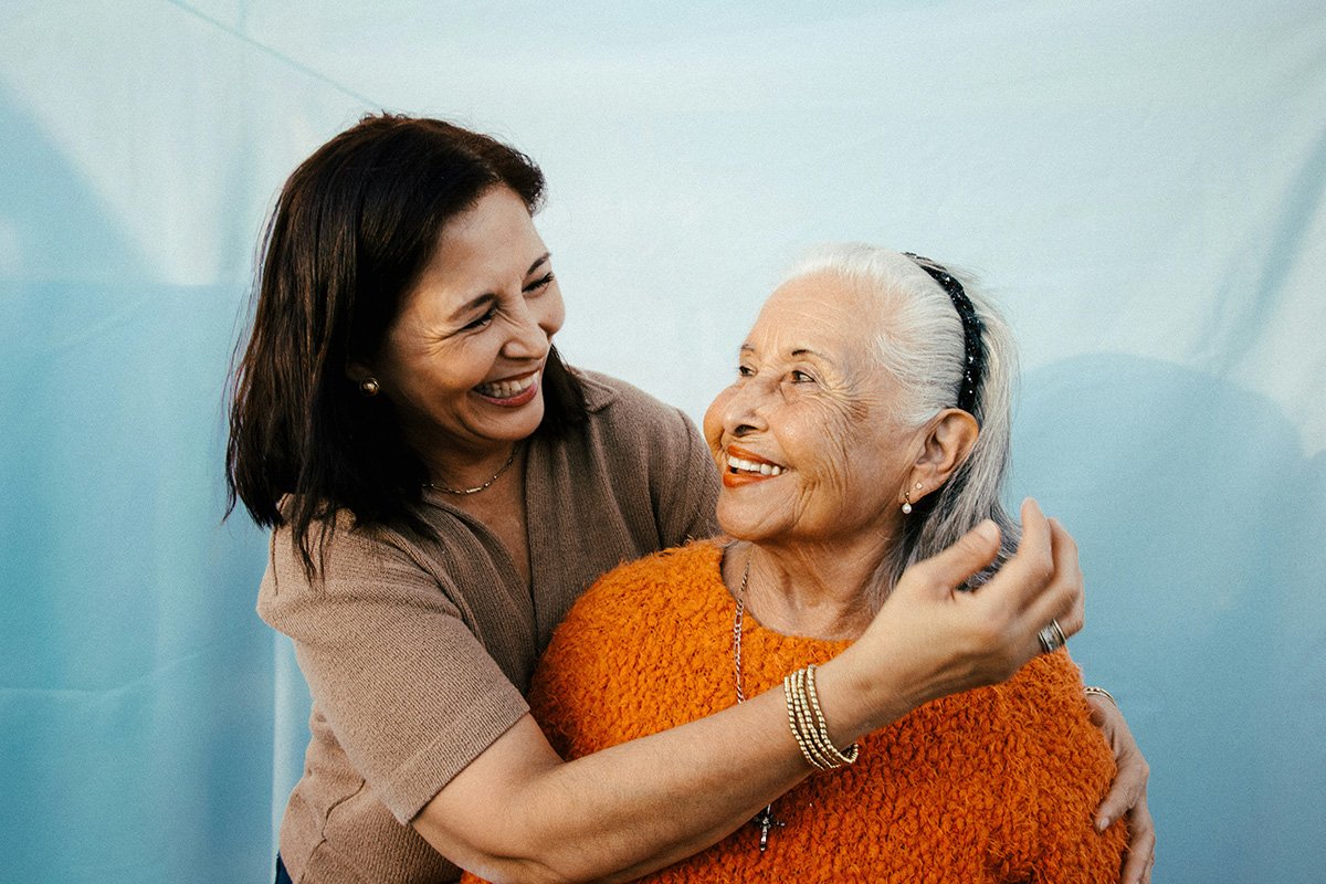 An adult hispanic woman with a brown hair smiles and embraces and elderly woman sith silver hair and an orange sweater. They are looking at each other and smiling.