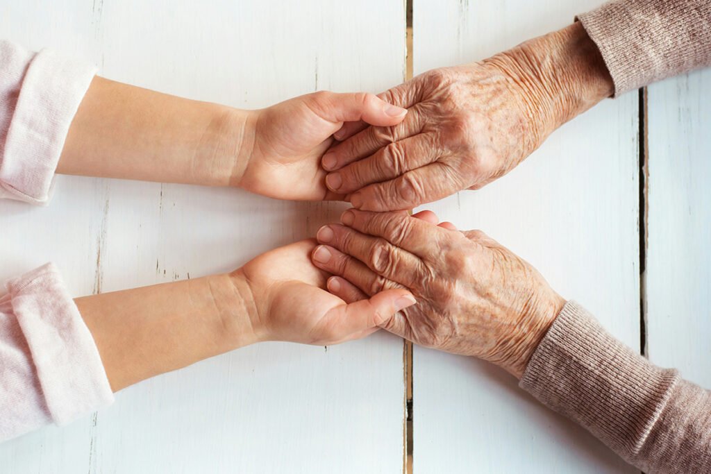 A young persons hands holding onto an elderly person’s hand across a wooden table.