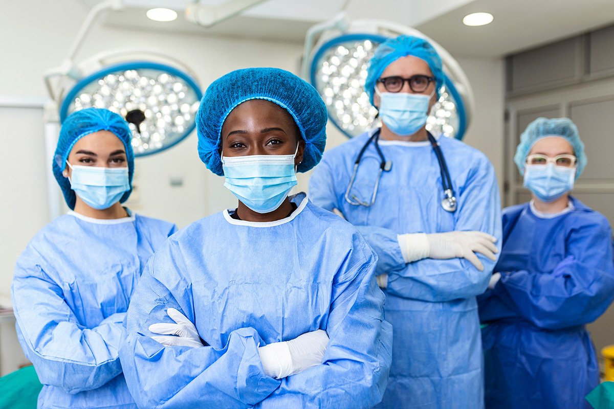 A team of diverse healthcare providers stand in an operation room wearing blue personal protective coverings and face masks as they look into the camera.