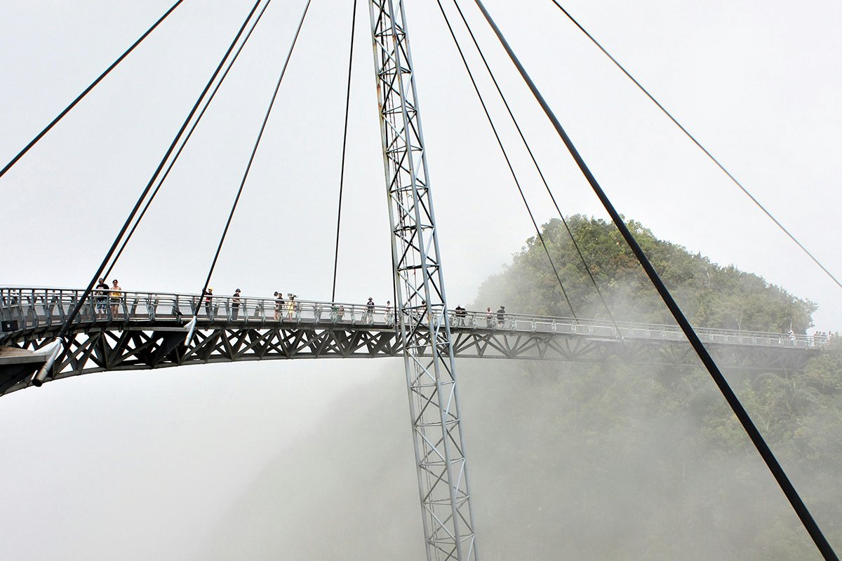 A very high cable bridge connects two separated pieces of land, with people walking from one side to the other. Langkawi Island, Kedah, Malaysia.