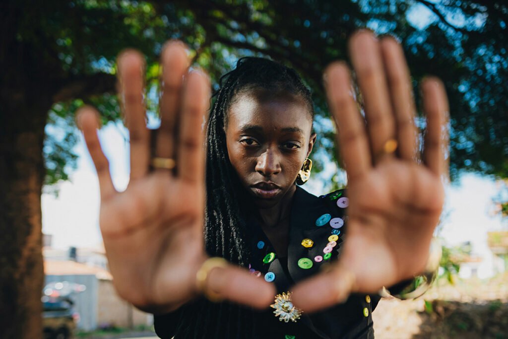 A Black woman with long braids stands under a tree, pushing her hands towards the camera, framing her own face with her hands.