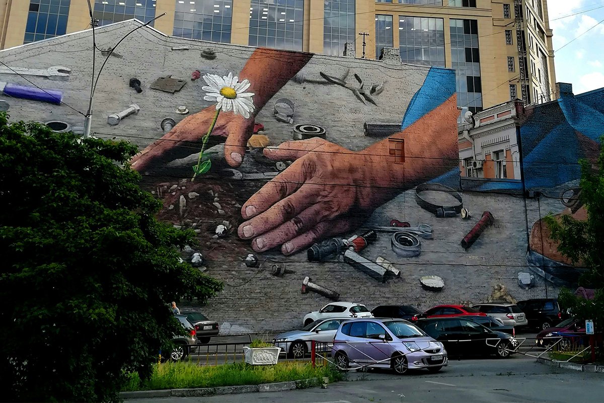 A mural painted on a building’s side depicting hands gathering soil around a sprouted daisy, with many work tools scattered around.