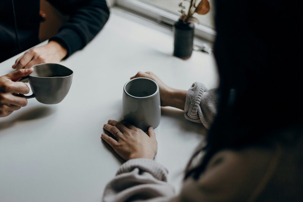 An over-the shoulder perspective of two people sitting at a table holding cups of coffee in their hands.