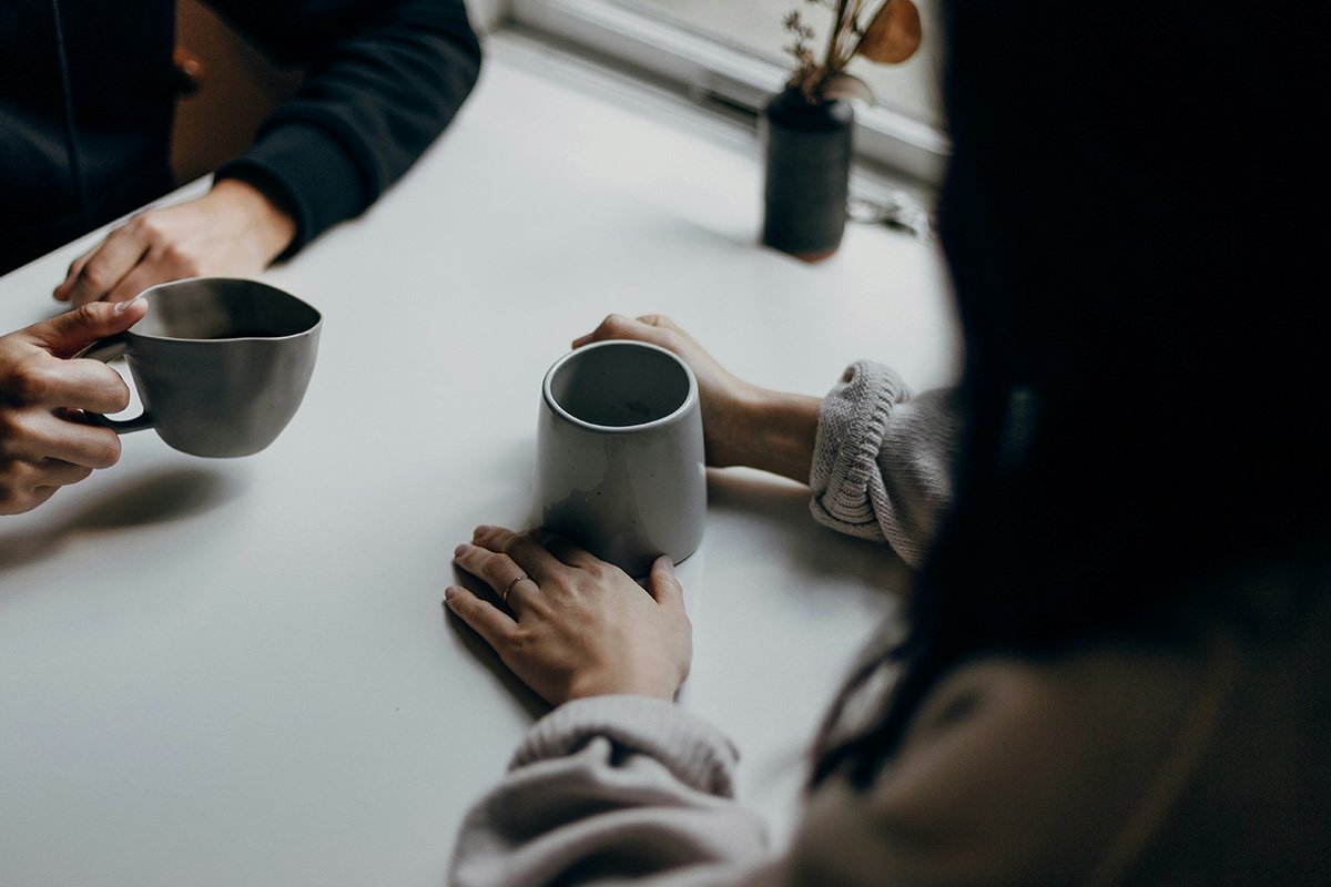 An over-the shoulder perspective of two people sitting at a table holding cups of coffee in their hands.