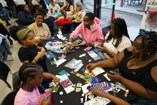 Several African American community members sit together at a table covered in squares of fabric to be used in a community quilt.