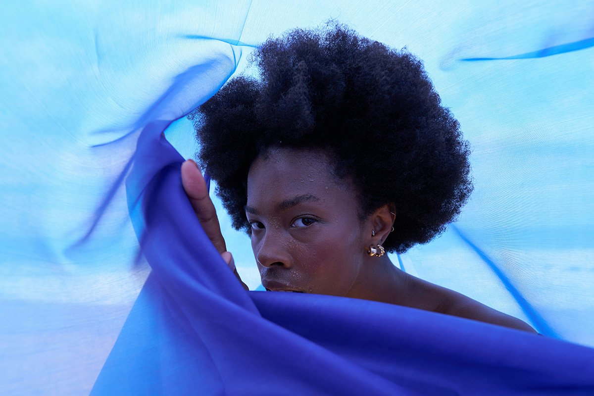 A Black woman looks intently into the camera, while holding a blue translucent scarf that envelopes the camera’s view, creating a tunnel around her.