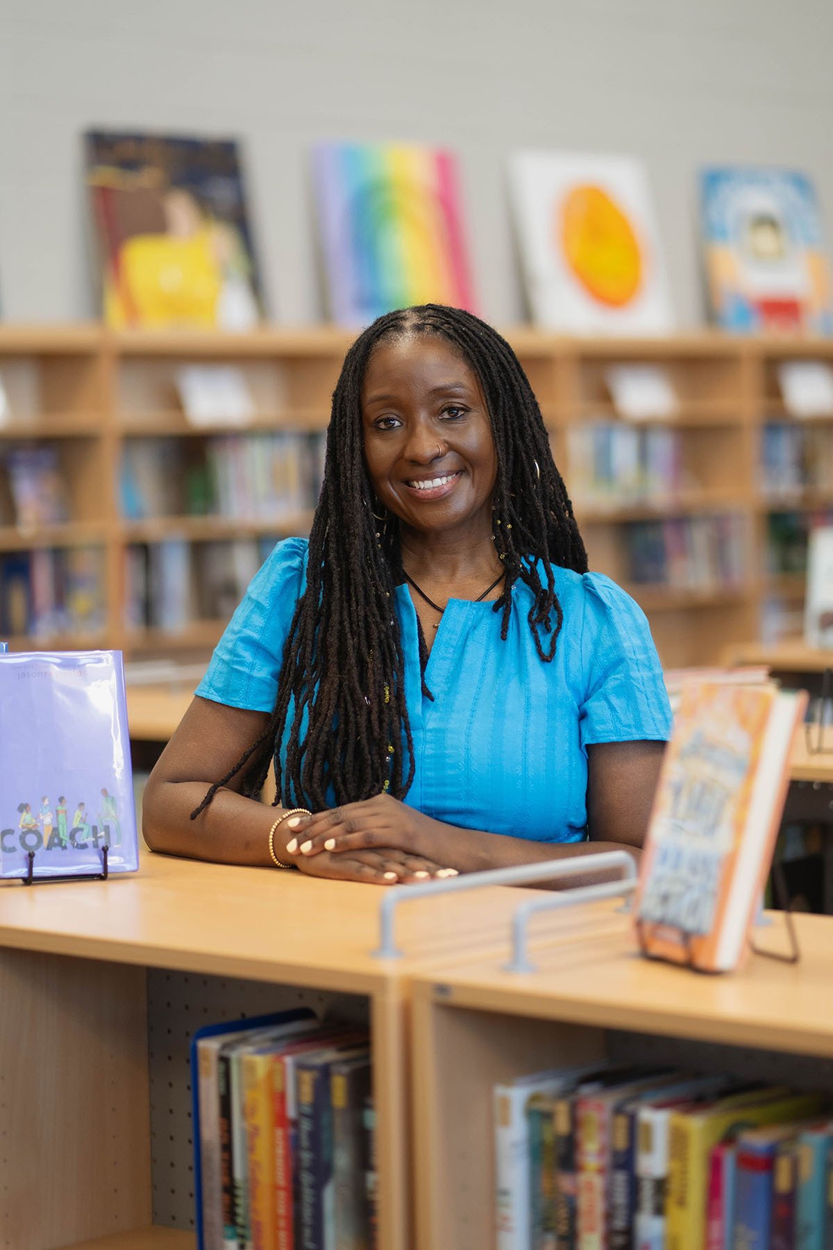 Cassie Owens Moore smiles while posing in a school library, with books displayed around her and bookshelves in the background.