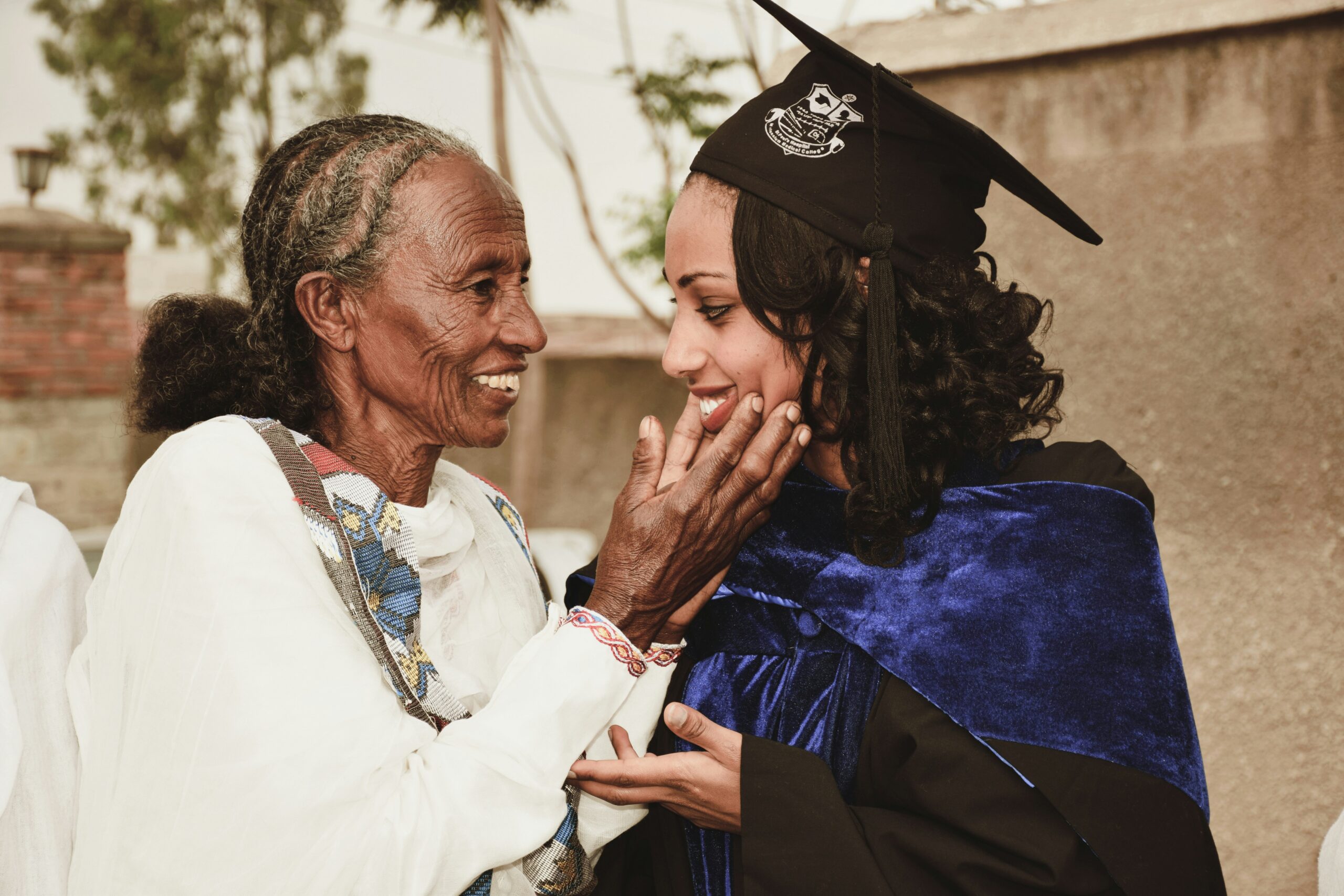 An elder cradles the face of a young woman in a graduation hat and robe