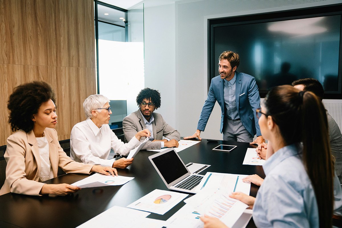 A board meeting takes place with a diverse group of board members sitting around a conference table in an office. There are papers with charts spread around the table.