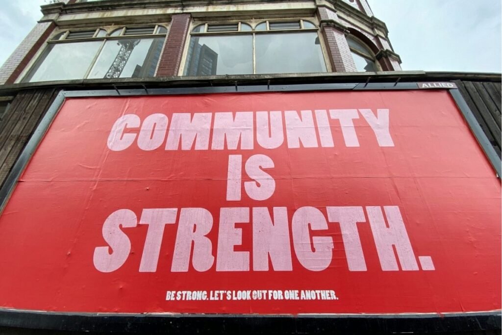 A large red banner on the side of a building reads "Community Is Strength. Be Strong. Let's Look Out For One Another.