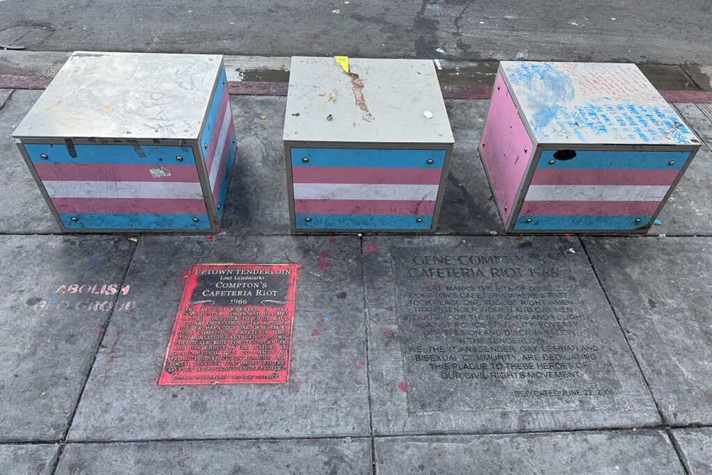 Two plaques commemorating Compton’s Cafeteria Riot on the sidewalk in the northwest corner of the intersection of Turk St and Taylor St in San Francisco. Behind the plaques are boxes painted with the trans flag.