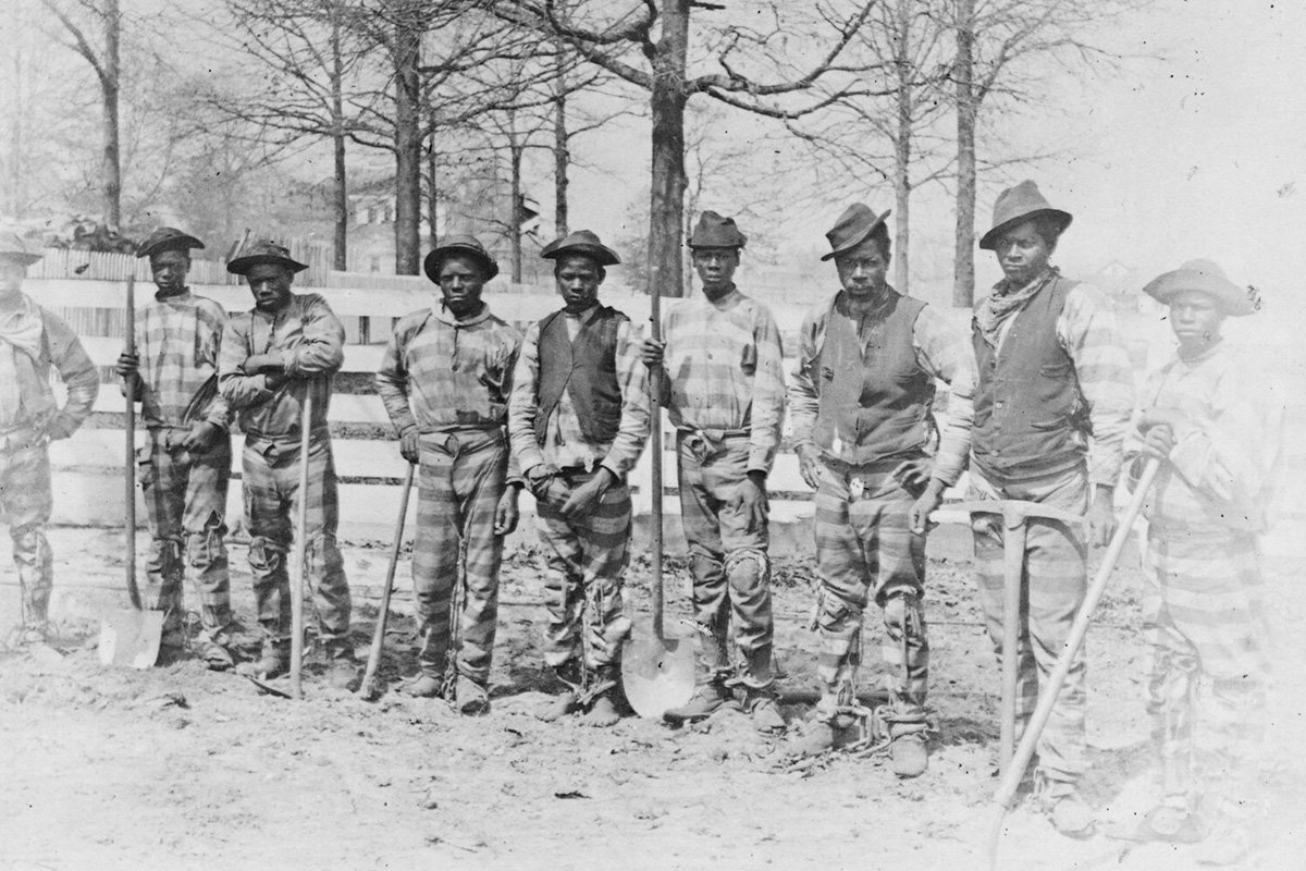 An antique black and white picture of a chain gang, Black men wearing striped clothing and chained together standing in a line. Thomasville, Georgia. 1884.