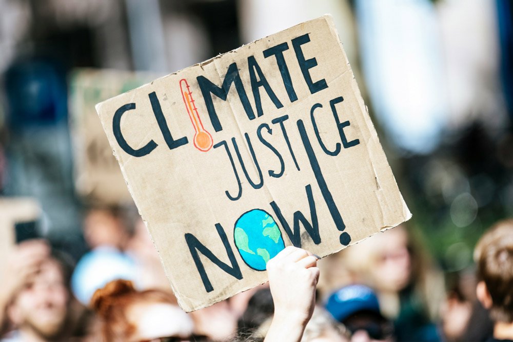 A hand-painted cardboard sign reading "Climate Justice Now" held above a crowd at a protest, with a thermometer and globe illustration drawn into the lettering.