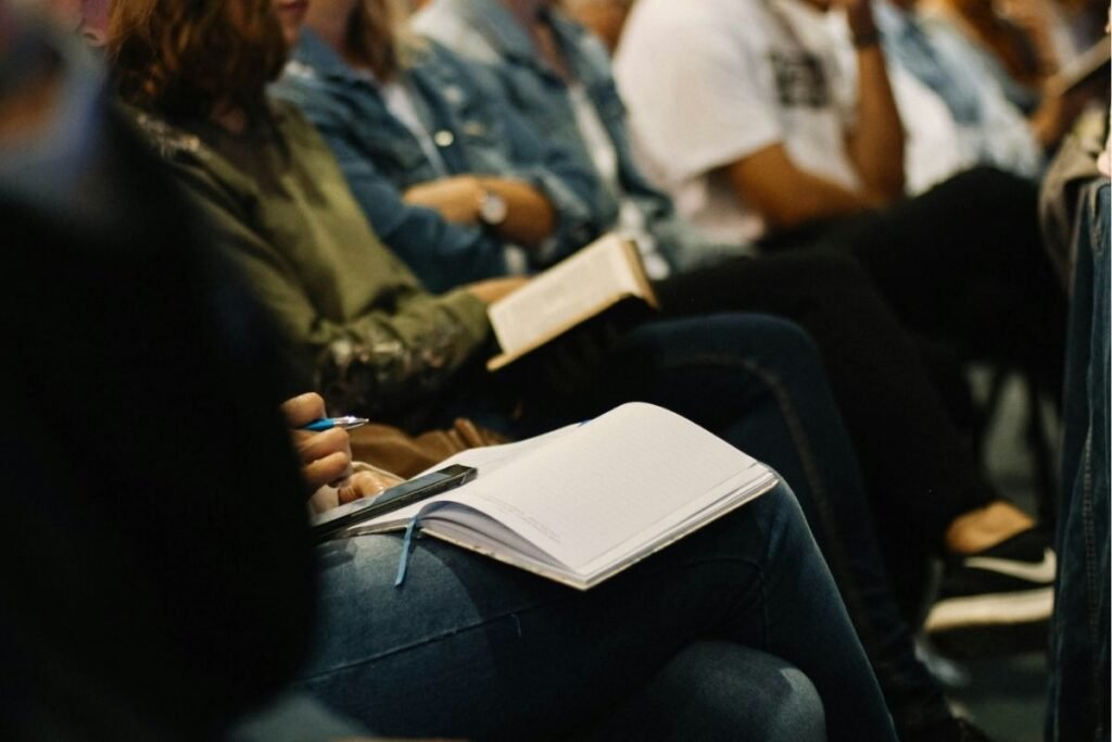 Community members seated at a public meeting, one attendee holding an open notebook and pen on their lap, ready to take notes.
