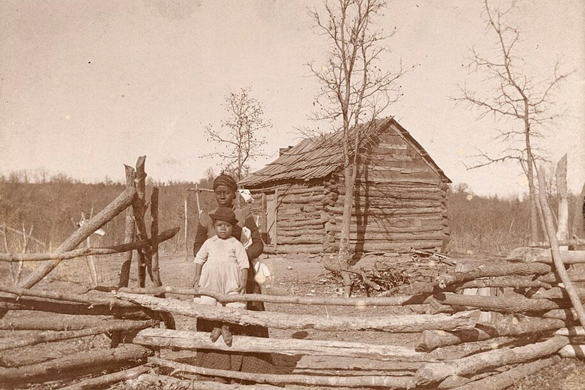 An antique sepia photograph depicting a Creek Freedmen mother and child standing outside of a log house in a field.