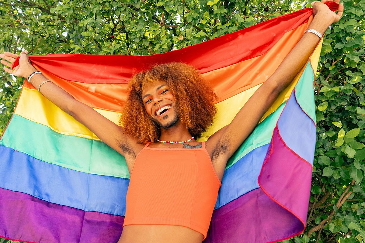 A person with red coily hair, a crop top, and a beard holds a rainbow flag behind them. In the background, there are trees.