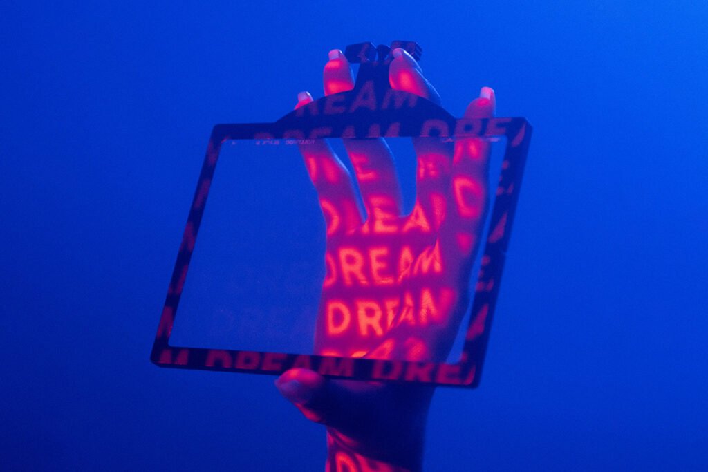 A woman’s hand in a dimly lit room, holding up a frame. The word “Dream” is projected onto her hand repeatedly.